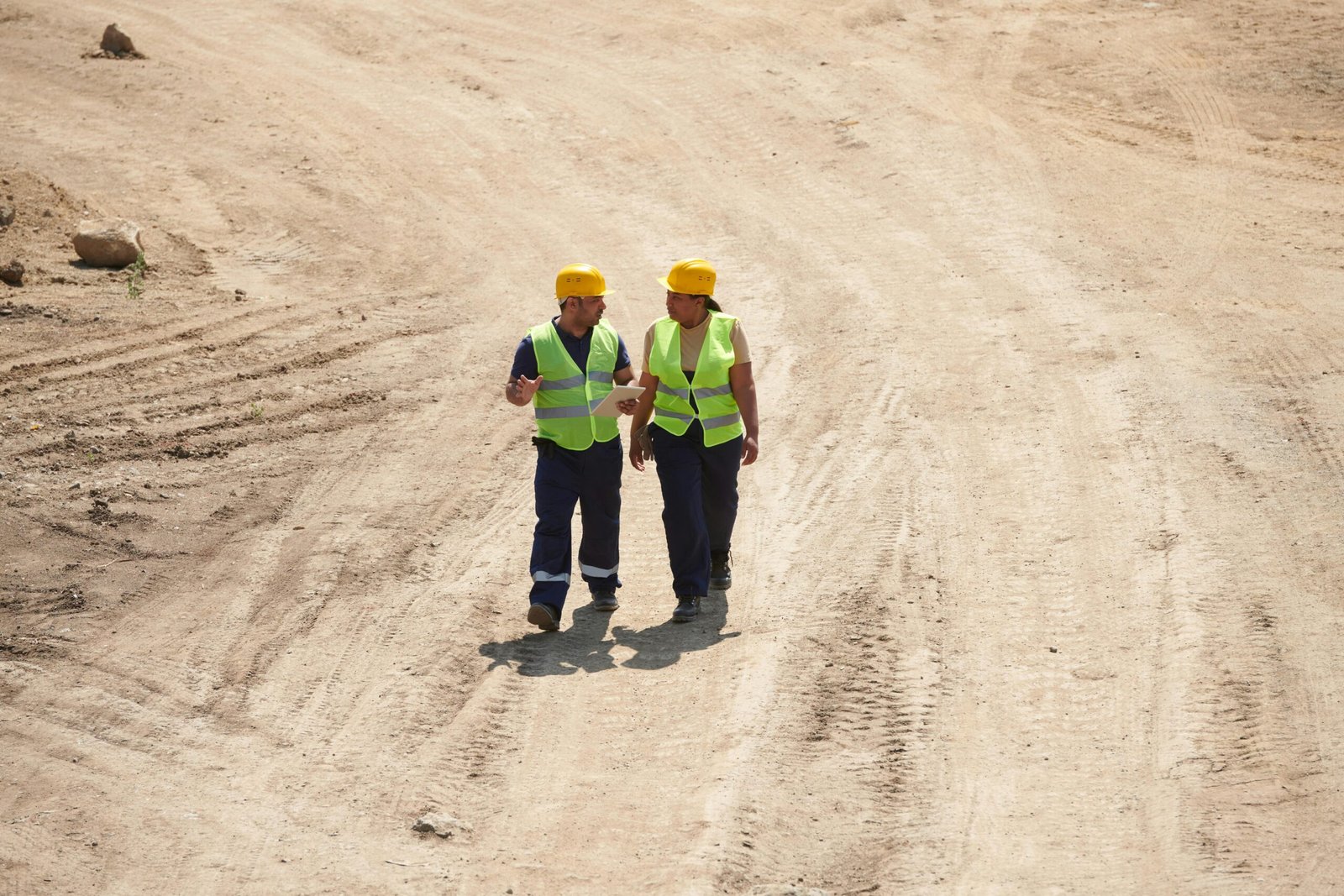 Two construction workers with safety vests and helmets on a construction site discussing plans.